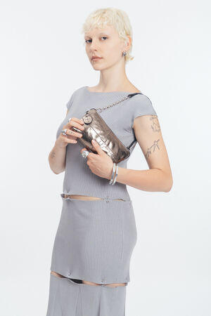 Person mit kurzen blonden Haaren in einem hellgrauen gerippten Kleid hält eine INA KENT Metallic-Handtasche mit einem Kettenriemen vor einem schlichten weißen Hintergrund.