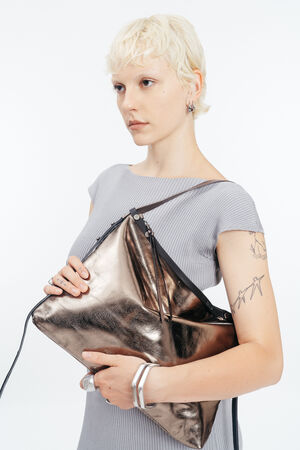 Eine Person mit kurzen blonden Haaren und einer Vogeltätowierung auf dem Arm hält eine große silberne INA KENT-Handtasche, während sie ein geripptes graues Kleid, silberne Ringe und Armbänder vor einem schlichten Hintergrund trägt.