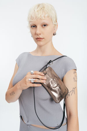 Eine Person mit kurzen blonden Haaren und sichtbaren Tätowierungen trägt ein geripptes graues Oberteil und hält eine kleine INA KENT Metallic-Crossbody-Tasche vor einem schlichten weißen Hintergrund.
