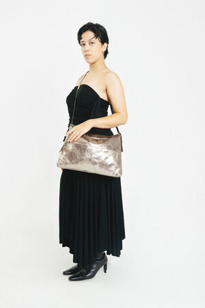 A person wearing a strapless black dress and black boots stands against a white background, holding an INA KENT metallic shoulder bag.