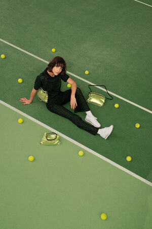 A person in a dark outfit and white sneakers sits on a green tennis court surrounded by yellow tennis balls and sleek metallic INA KENT bags.