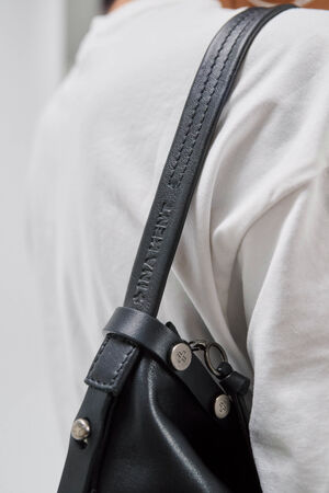 Nahaufnahme einer Person in einem weißen Hemd, die eine schwarze Ledertasche von INA KENT über der Schulter trägt; der Riemen ist mit geprägtem Text und Metallverschlüssen versehen.
