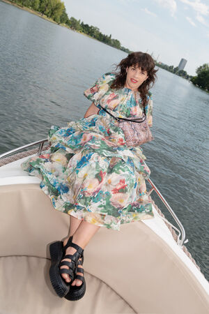A person in a floral dress and black sandals sits on the edge of a boat, holding an INA KENT patterned bag, with water and trees in the background.