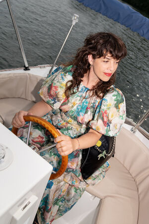 A woman in a floral dress sits at the helm of a boat, steering with one hand and looking to the side. An INA KENT bag rests nearby, while water and part of the boat's canopy are visible in the background.