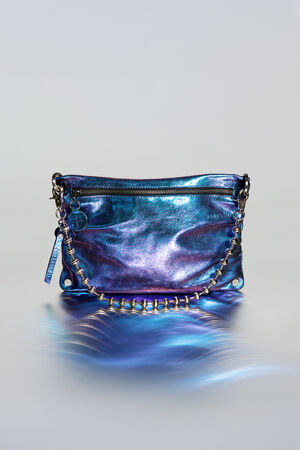 Metallisch blau schillernde Handtasche von INA KENT mit Reißverschluss, dekorativem Kettenhenkel und rechteckiger Form vor einem hellen Hintergrund mit Farbverlauf.