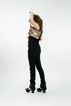 A person in black clothing and platform sandals stands against a plain background, holding an INA KENT metallic handbag over their shoulder with their face turned away.