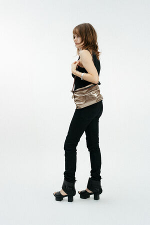 A person in black clothing and heeled sandals stands sideways, holding an INA KENT metallic crossbody bag against a plain white background.
