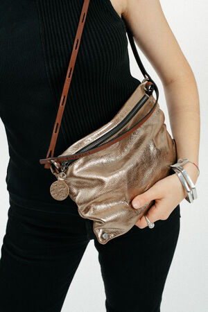 A person wearing a black sleeveless top and pants holds an INA KENT metallic bronze crossbody bag with a circular keychain and brown strap.