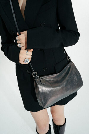 Eine Person, die ein schwarzes Blazerkleid und Ringe trägt, hält eine INA KENT-Metallic-Schultertasche und ein silbernes Accessoire, wobei kniehohe schwarze Stiefel zu sehen sind.