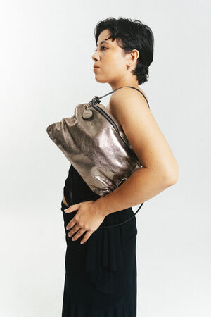 Person in a black outfit standing sideways, holding a large metallic INA KENT purse under their arm against a plain white background.
