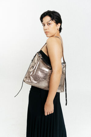 Eine Person mit kurzen dunklen Haaren und einem schwarzen Kleid steht seitlich und trägt eine INA KENT Metallic-Umhängetasche mit Reißverschluss und Riemen vor einem schlichten weißen Hintergrund.