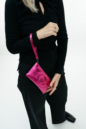 Eine schwarz gekleidete Person hält eine INA KENT Metallic-Pink-Armbandtasche in ihrer linken Hand vor einem weißen Hintergrund.