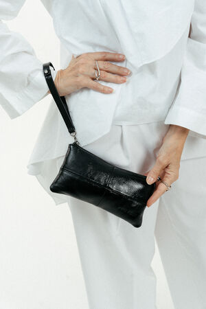 A person in a white outfit holds an INA KENT black leather wristlet purse against their hip.