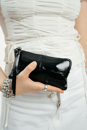 Eine Person, die ein weißes Rüschenoberteil und weiße Jeans trägt, hält eine kleine schwarze INA KENT-Armbandtasche aus Lackleder.