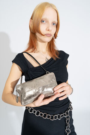 Person mit heller Haut und glattem, schulterlangem Haar hält eine silberne Metallic-Handtasche von INA KENT, während sie ein schwarzes Kleid und einen klobigen Kettengürtel trägt.