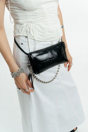 A person wearing a white outfit with a black INA KENT patent leather crossbody bag and silver accessories, including rings and a beaded bracelet.