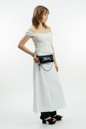 A woman stands in profile wearing an off-shoulder white top, long white skirt, black platform sandals, and an INA KENT black belt bag with a pearl chain.