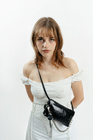 A woman with shoulder-length brown hair wears an off-the-shoulder white dress and an INA KENT black crossbody bag, standing against a plain white background.