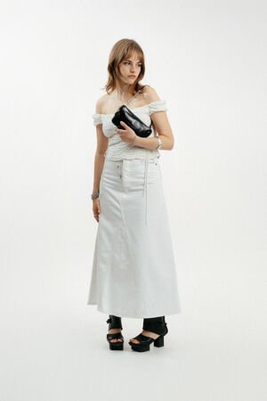A woman wearing an off-shoulder white top and long white skirt, holding an INA KENT black clutch, and wearing black platform sandals, stands against a plain white background.