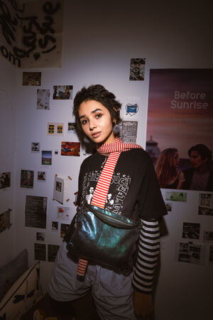 Eine junge Person steht in einem geschmückten Raum, trägt einen gestreiften Schal, ein grafisches T-Shirt und eine metallene INA KENT-Tasche, während an der Wand hinter ihr Fotos und ein "Before Sunrise"-Poster hängen.