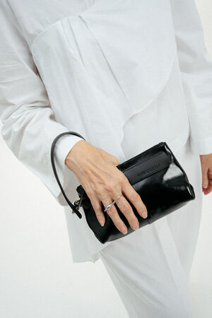 Eine weiß gekleidete Person hält eine kleine schwarze Lederhandtasche von INA KENT mit einer Handschlaufe in ihrer linken Hand.