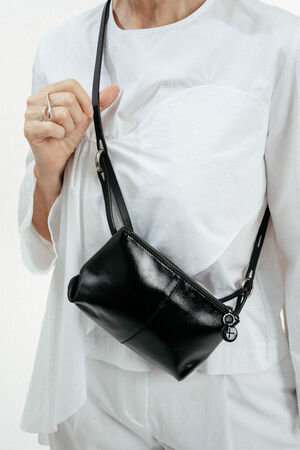 Eine weiß gekleidete Person trägt eine kleine schwarze INA KENT-Lederkreuztasche mit silbernem Reißverschluss und Ringdetails.