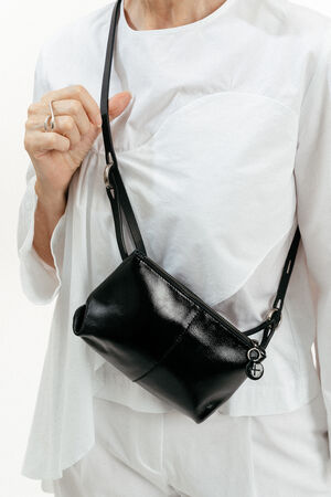 Eine weiß gekleidete Person hält eine kleine schwarze INA KENT-Ledertasche mit einem silbernen Anhänger in der Hand, deren Riemen sie sich über die Schulter gelegt hat.