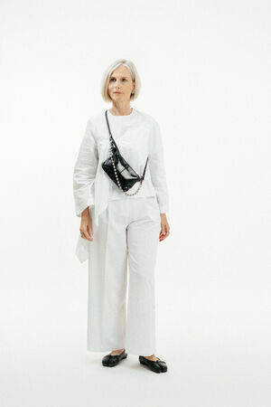 Eine ältere Frau mit kurzen grauen Haaren steht vor einem weißen Hintergrund. Sie trägt ein weißes Outfit, schwarze Schuhe und eine schwarze INA KENT Crossbody Bag.