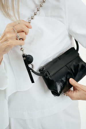 Person wearing a white outfit holds an INA KENT black leather handbag with a metal chain strap and decorative rings, featuring a unique ring on one finger.