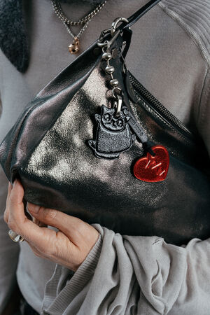 A person holds an INA KENT metallic handbag adorned with a cat keychain and a red heart keychain attached to the strap.