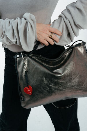 A person wearing a light gray top and black pants holds an INA KENT metallic handbag adorned with a red heart-shaped keychain.