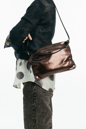 Person, die einen durchsichtigen schwarzen Pullover über einem gepunkteten Hemd trägt und eine INA KENT Metallic-Bronze-Umhängetasche in der Hand hält; dunkle Jeans sind zu sehen.