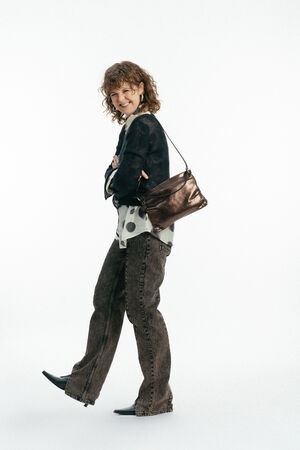 Person mit lockigem Haar, die einen schwarzen Pullover, ein gepunktetes Hemd, braune Jeans und schwarze Stiefel trägt und eine INA KENT Umhängetasche in Bronze-Metallic mit sich führt, posiert vor einem schlichten weißen Hintergrund.