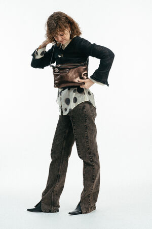 Person mit lockigem Haar, die eine Brille, eine schwarze Jacke, ein gepunktetes Hemd, braune Jeans und schwarze Schuhe trägt, hält eine INA KENT Metallic-Umhängetasche. Weißer Hintergrund.