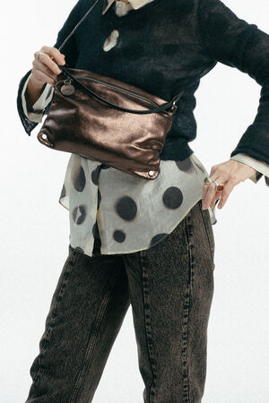 Eine Person, die eine durchsichtige gepunktete Bluse, einen dunklen Pullover und Jeans trägt, posiert mit einer metallischen INA KENT Crossbody Bag.