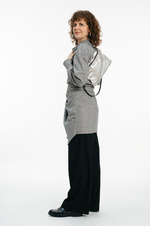 Eine Frau mit lockigem Haar steht seitlich und trägt ein graues langärmeliges Oberteil, eine schwarze Hose mit weitem Bein, schwarze Schuhe und einen silberfarbenen INA KENT-Rucksack über einer Schulter.