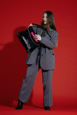 A woman in a gray suit stands against a red background, looking into her INA KENT black tote bag while holding a shiny magenta item.