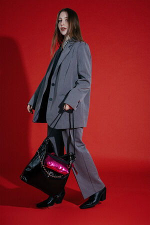 A person in a gray oversized suit and black boots stands against a red background, holding an INA KENT large black bag with a shiny pink pouch attached.