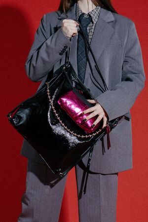 Person in a gray suit holding a large black INA KENT patent leather bag and a shiny pink clutch against a red background.