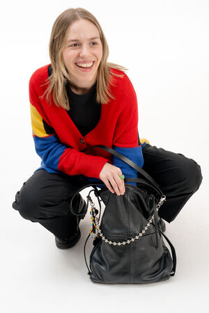 Person in a red, blue, and yellow cardigan, black pants, and black shoes, squatting and smiling next to a large INA KENT black handbag with chain detail, on a white background.