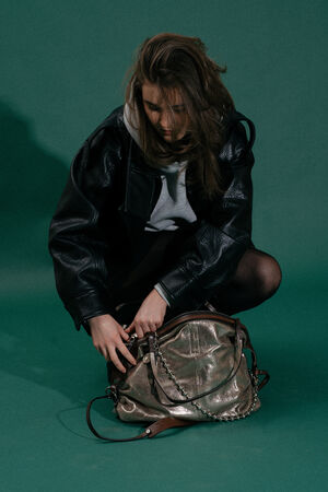 A person crouches on a green background, wearing a black leather jacket and gray hoodie, looking down while searching through an INA KENT metallic handbag.