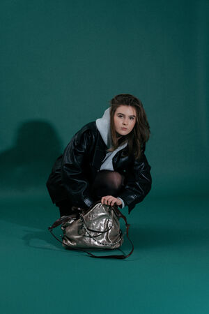 A young woman in a black leather jacket and grey hoodie crouches on a green background, holding an INA KENT metallic handbag on the floor.