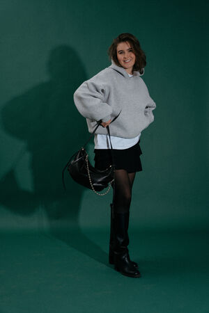 A person stands on a green backdrop, wearing a gray sweatshirt, white shirt, black skirt, black tights, knee-high boots, and holding an INA KENT black handbag.
