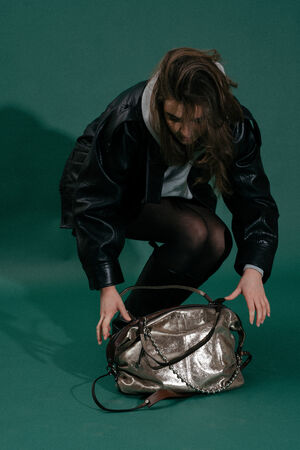 A person in a black jacket and tights is crouching down, reaching toward an INA KENT metallic handbag on a green background.