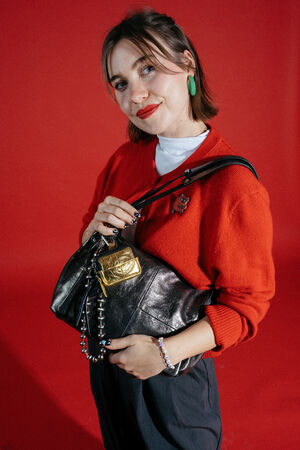 Woman in a red sweater with green earrings poses holding an INA KENT black handbag with a gold accent against a solid red background.