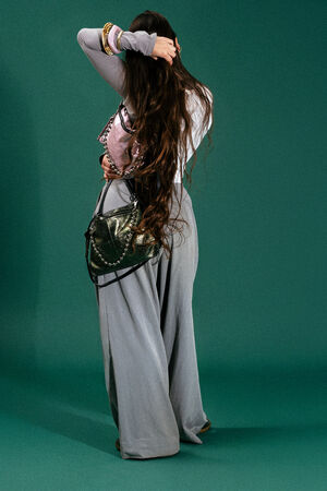 A person with long brown hair stands against a green background, wearing wide gray pants, a light top, bracelets, and carrying an INA KENT dark green handbag. Their face is turned away.