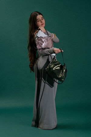 Woman with long hair wearing a gray outfit poses against a green background, holding an INA KENT metallic purple purse on her shoulder and a shiny dark green handbag in her hand.