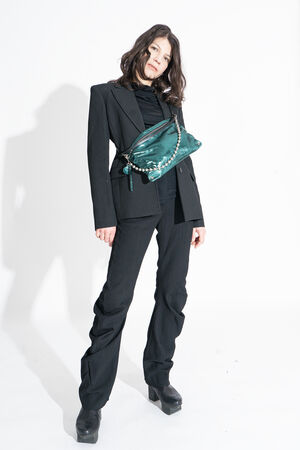 A woman in a black suit and platform shoes stands against a white background, wearing an INA KENT green crossbody bag.