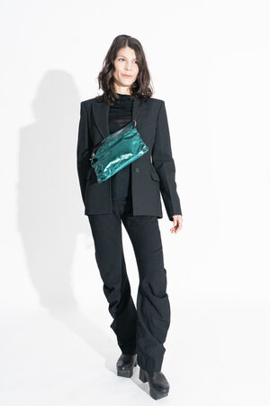 A woman in a black suit and platform shoes stands against a white background, wearing a shiny green INA KENT crossbody bag.