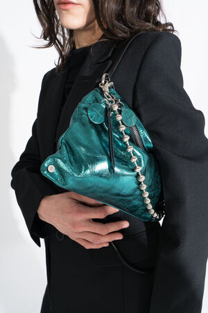 Person in a black jacket holding an INA KENT shiny teal metallic handbag with chain and zipper details against a plain white background.